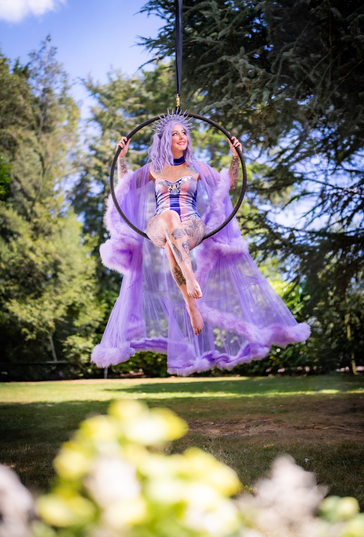 A performer in a flowing purple costume sits in an aerial hoop outdoors among trees.