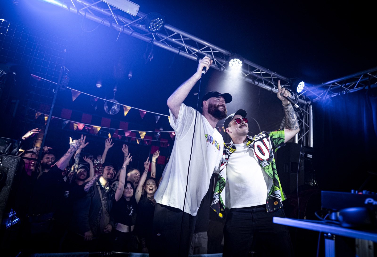 Two performers raise their microphones under blue stage lights, with a cheering crowd behind them.